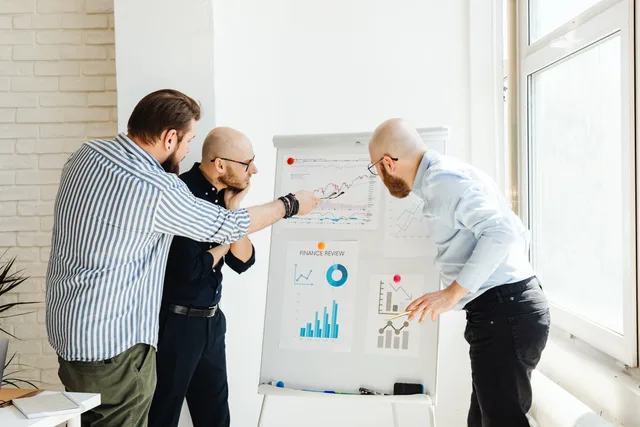 Three colleagues in a bright office discuss financial charts on a whiteboard. Investigating many leads but few accepted opportunities