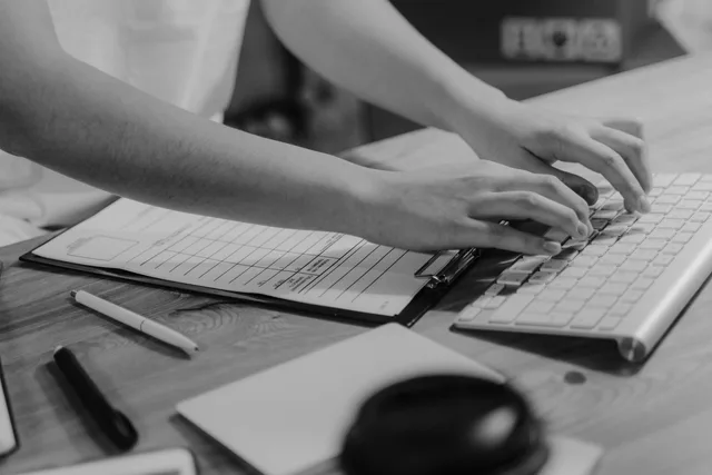 Black-and-white photo of hands typing on keyboard with clipboard on wooden desk. Lead generation data hygiene